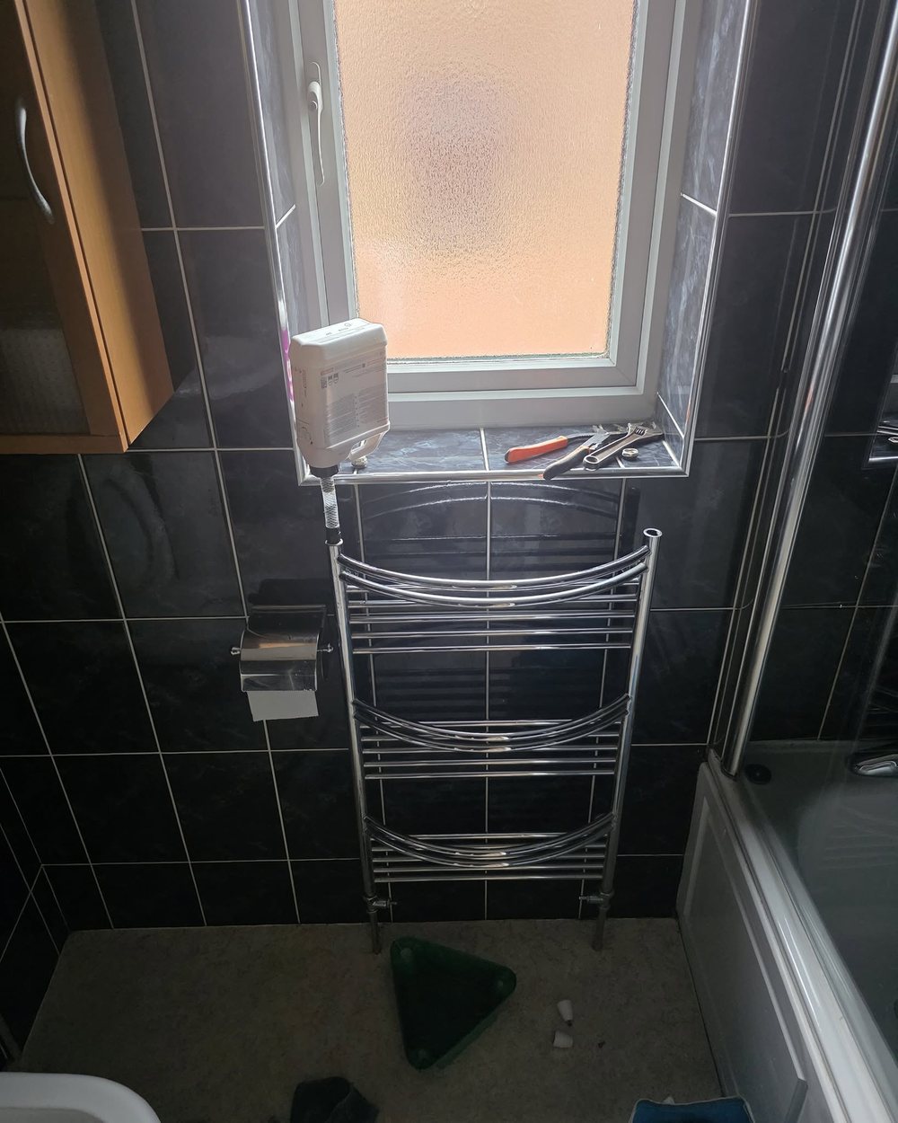 Chrome ladder heated towel rail installed against black tiled bathroom walls